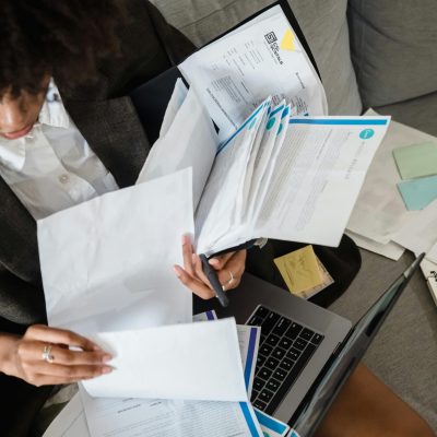A woman sitting on a couch organizing paperwork with a laptop in front of her.